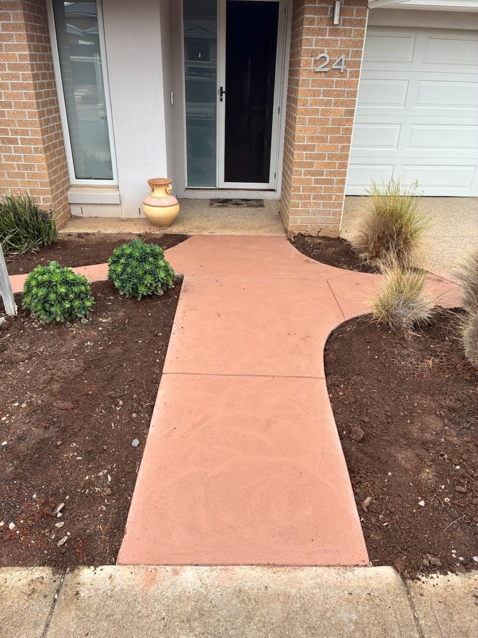 Red concrete walkway leading to a house entrance. Mulch and plants flank the path.