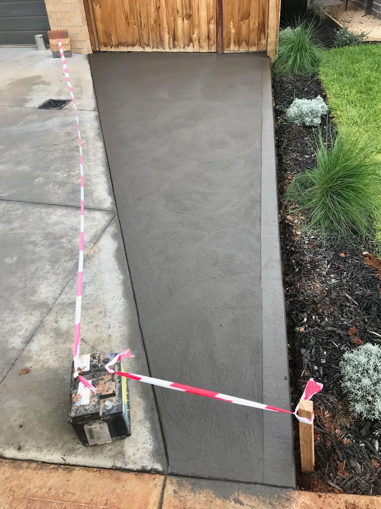 New concrete pathway leading to a wooden garage door. Caution tape surrounds the pathway; garden bed with plants on the right.