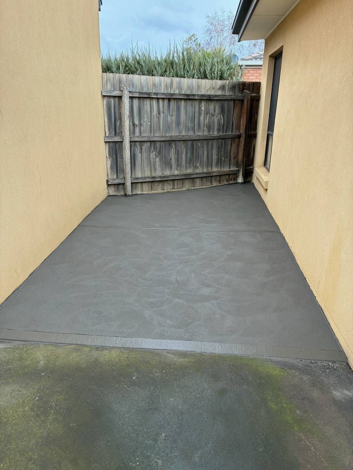 A freshly poured concrete patio between two beige walls leading to a wooden fence.