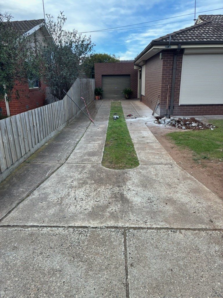 Concrete driveway with a strip of grass leading to a garage, flanked by a fence and a brick house.