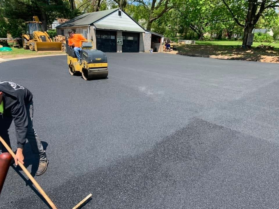 Workers paving a driveway with a roller machine; a worker rakes the fresh asphalt.