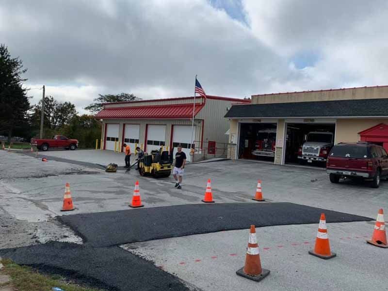 Asphalt paving in progress at a fire station. Workers use a roller machine on a fresh patch surrounded by safety cones; fire trucks are visible inside the bays.
