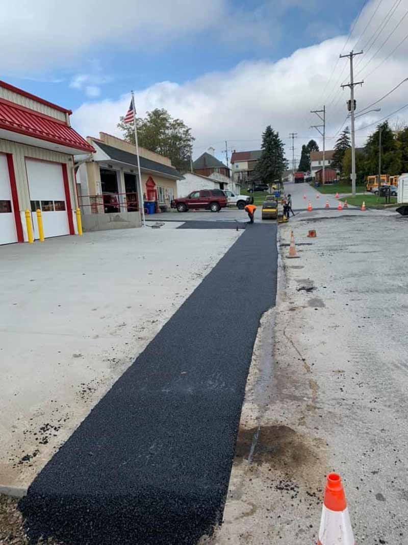 Asphalt paving in progress near a building with red doors. Workers and equipment visible on a street with traffic cones.