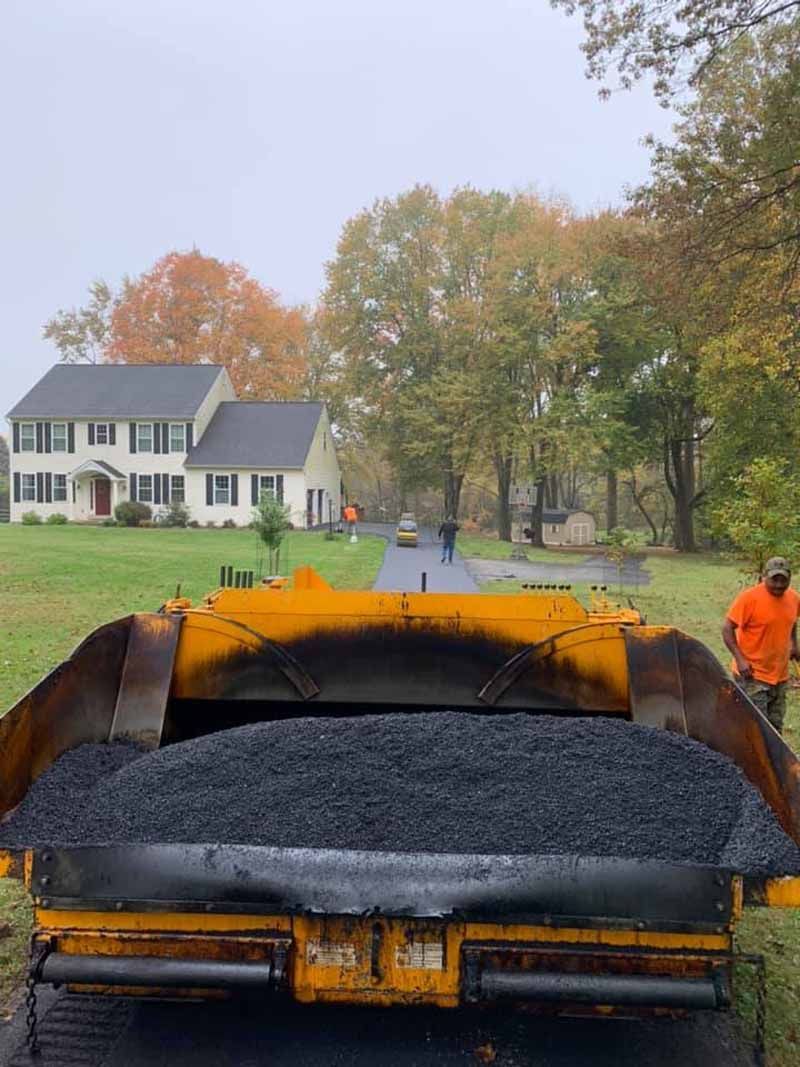 Asphalt paving a driveway in front of a house. A yellow asphalt spreader holds the blacktop. Two workers are present.
