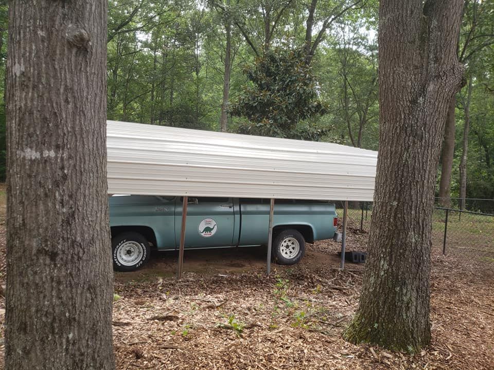 A blue truck is parked under a carport in the woods.