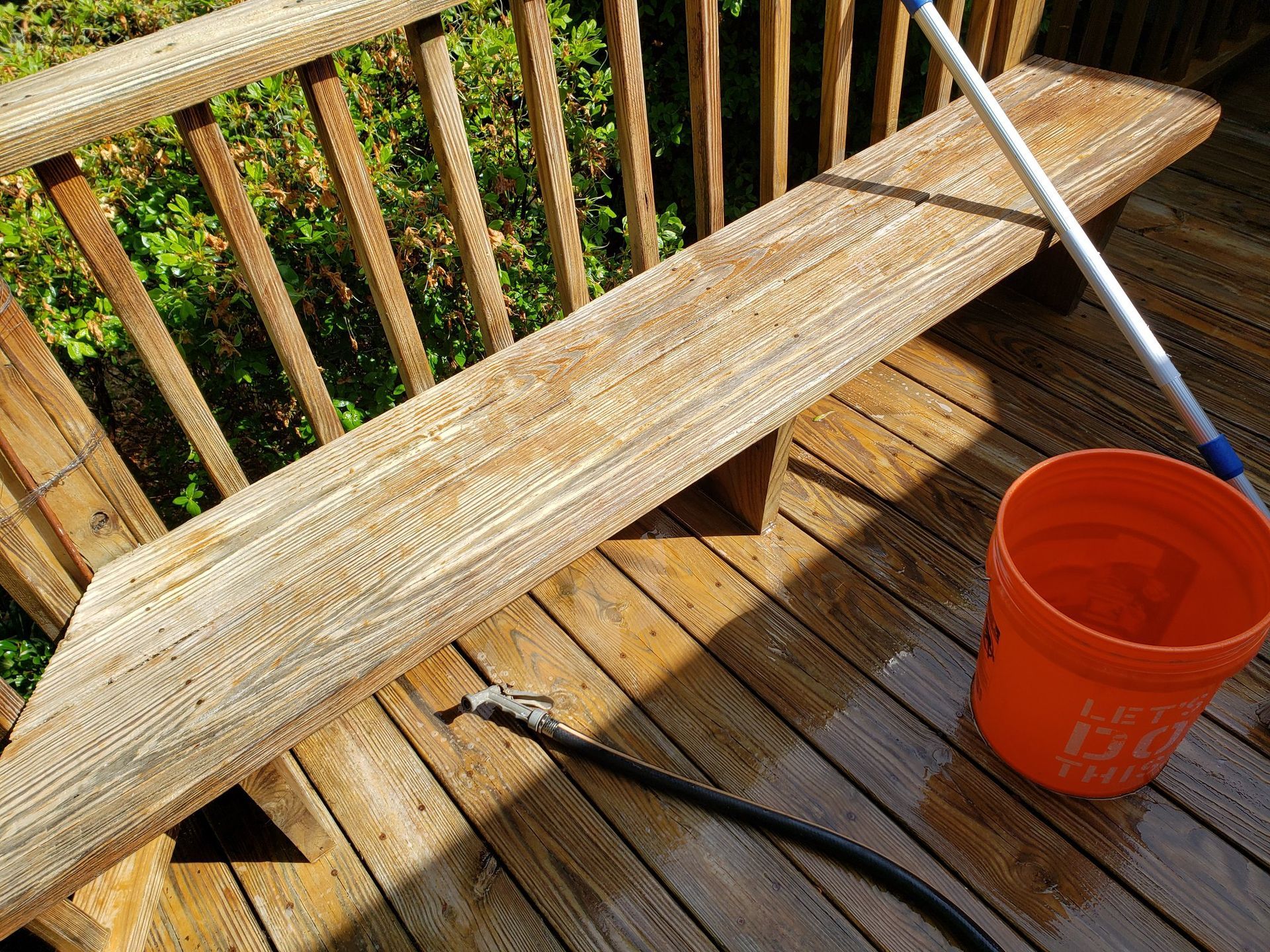 A wooden bench is being cleaned on a deck with a hose and bucket.