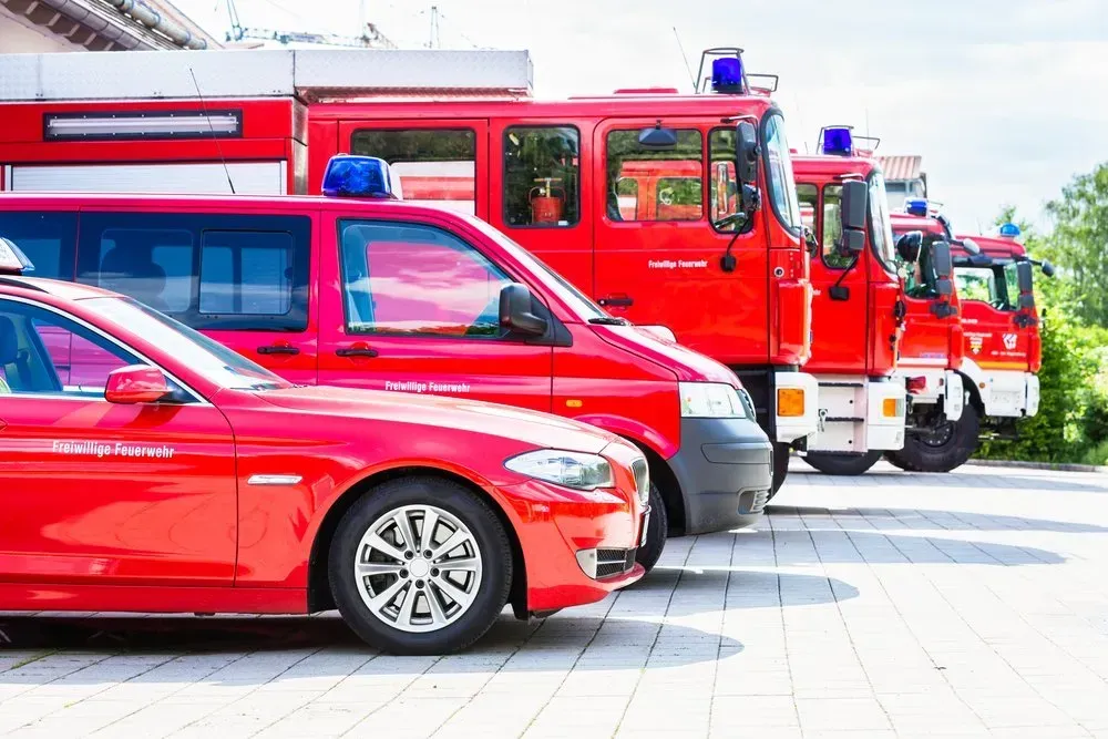 A row of red fire trucks and cars are parked in a parking lot.