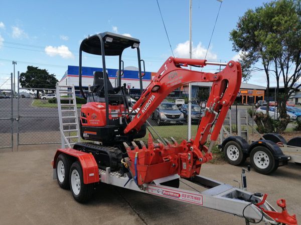 A small red excavator is on a trailer in a parking lot.