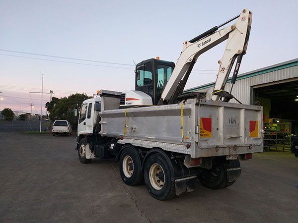 A bobcat excavator is attached to the back of a dump truck.