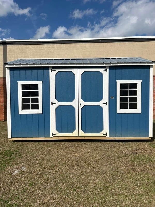 Blue shed with white trim, double doors, and windows, set against a building and blue sky.