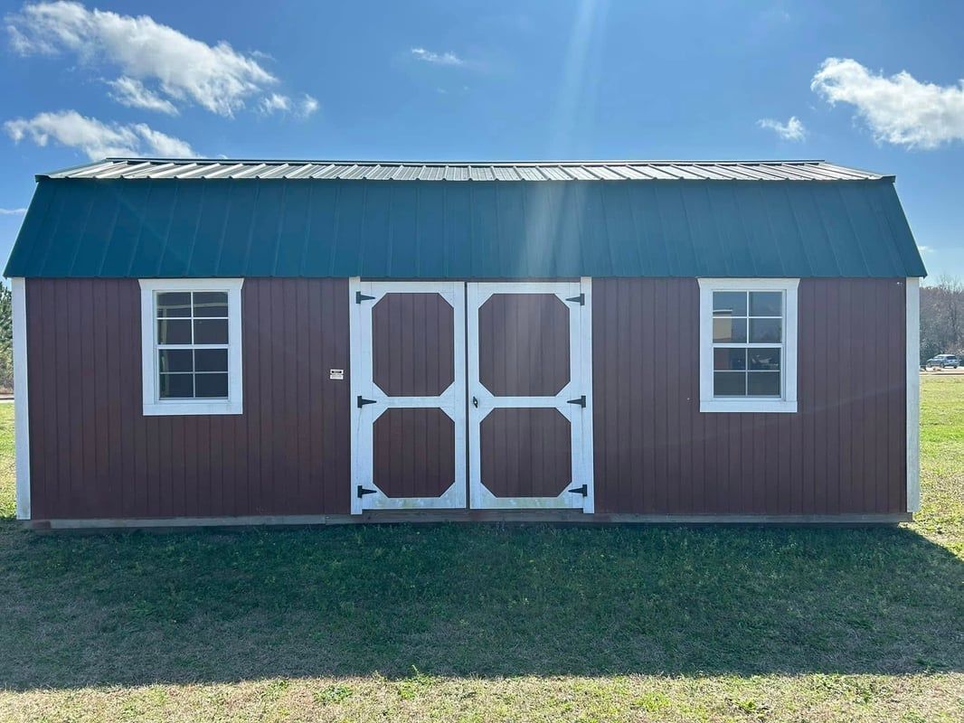 Brown barn with green roof, white doors and windows on grassy ground under a blue sky.