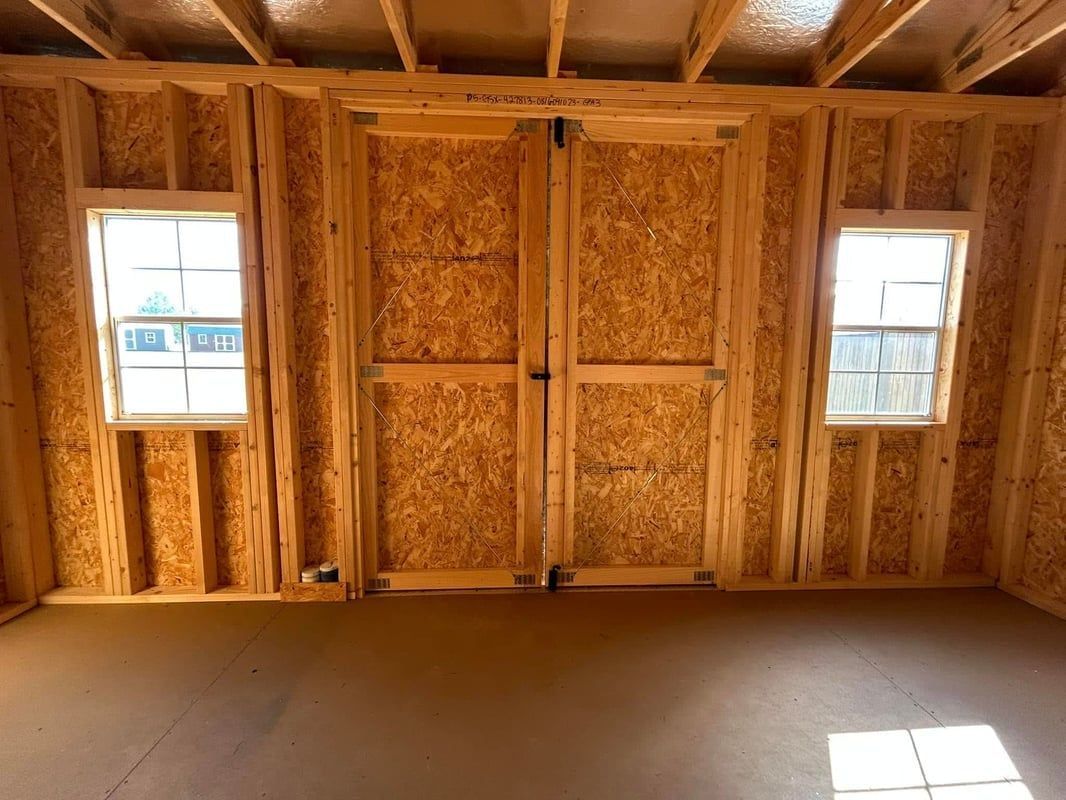 Interior of a wood-framed shed with two windows flanking a pair of wooden doors; unfinished floor and ceiling.