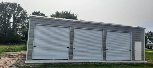 Three-bay garage with roll-up doors; gray siding; green lawn and trees in the background.