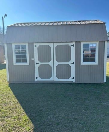 Tan storage shed with white-trimmed windows and double doors, sitting on grass under a blue sky.