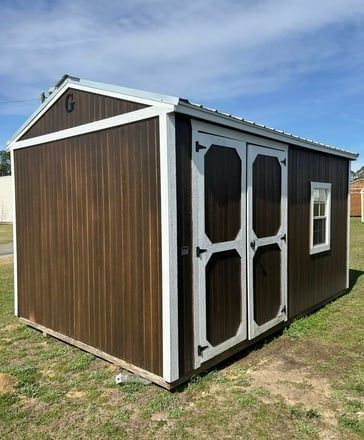 Brown shed with white trim and double doors on a grassy area under a blue sky.