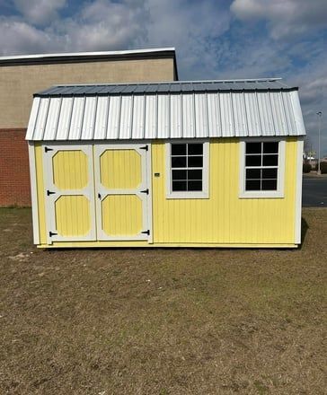 Yellow shed with white trim, doors, and two windows. Gray metal roof. Sits on grass.