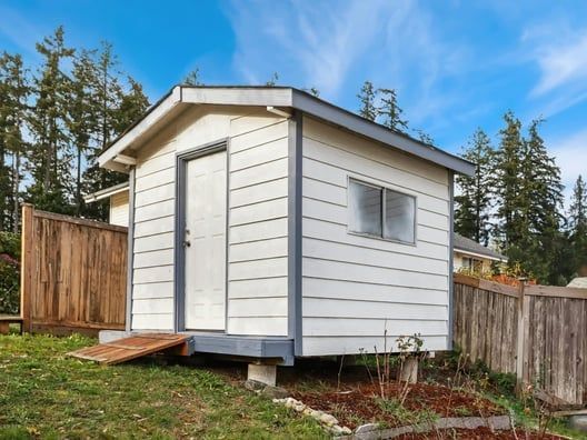 White shed with gray trim, door, window, and small ramp on grass lawn next to a wooden fence.