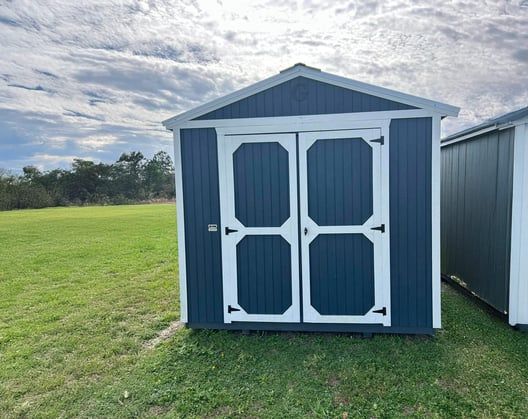 Blue storage shed with white trim and double doors on a grassy lawn under a cloudy sky.