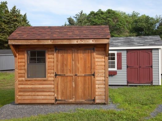Wooden shed with brown roof and doors, window to the left. Another shed with red doors and white trim to the right.