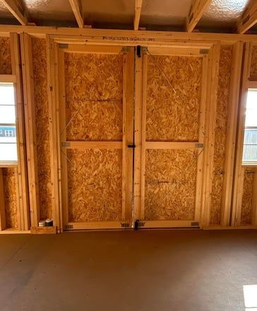 Interior of a wooden shed with double doors made of particleboard and two small windows.