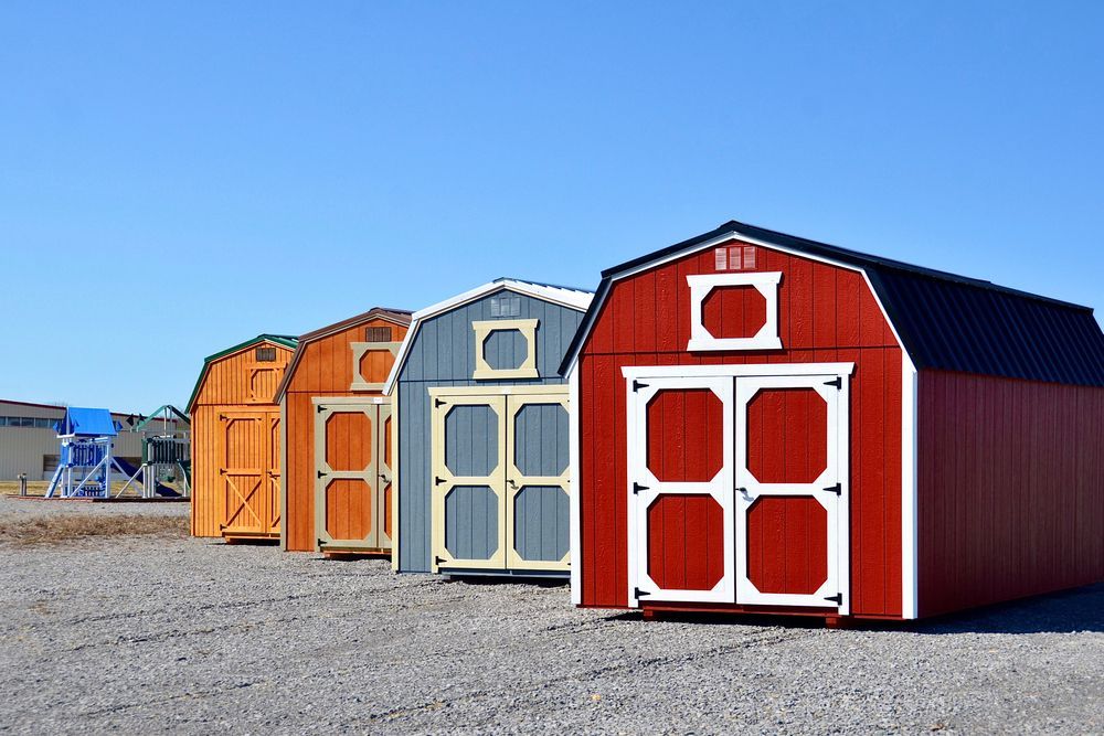 Four colorful sheds lined up in a row on a gravel lot under a clear blue sky.
