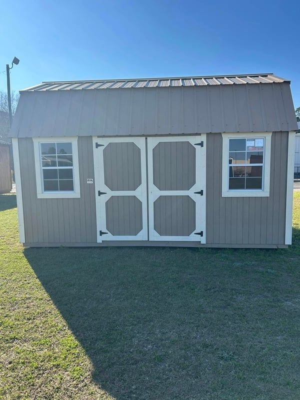 Tan shed with white-trimmed double doors and windows, sitting on green grass under a clear sky.