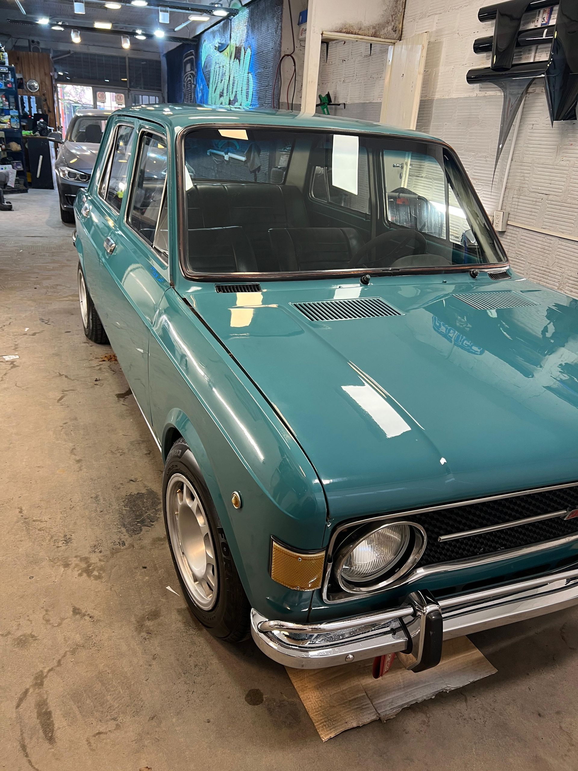Coche clásico verde azulado aparcado en un garaje. Detalles cromados, faros redondos y llantas deportivas.