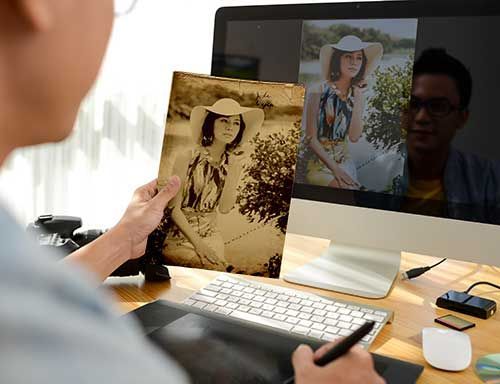 Photo editing image, man comparing physical photo he is restoring to the digital version he has edited on computer monitor