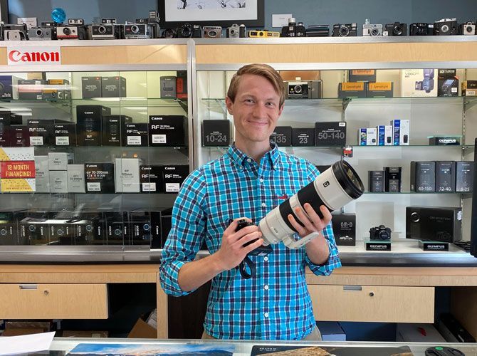 Employment opportunities image, image of young man in blue plaid dress shirt holding pro camera lens while standing in in front of product display case.