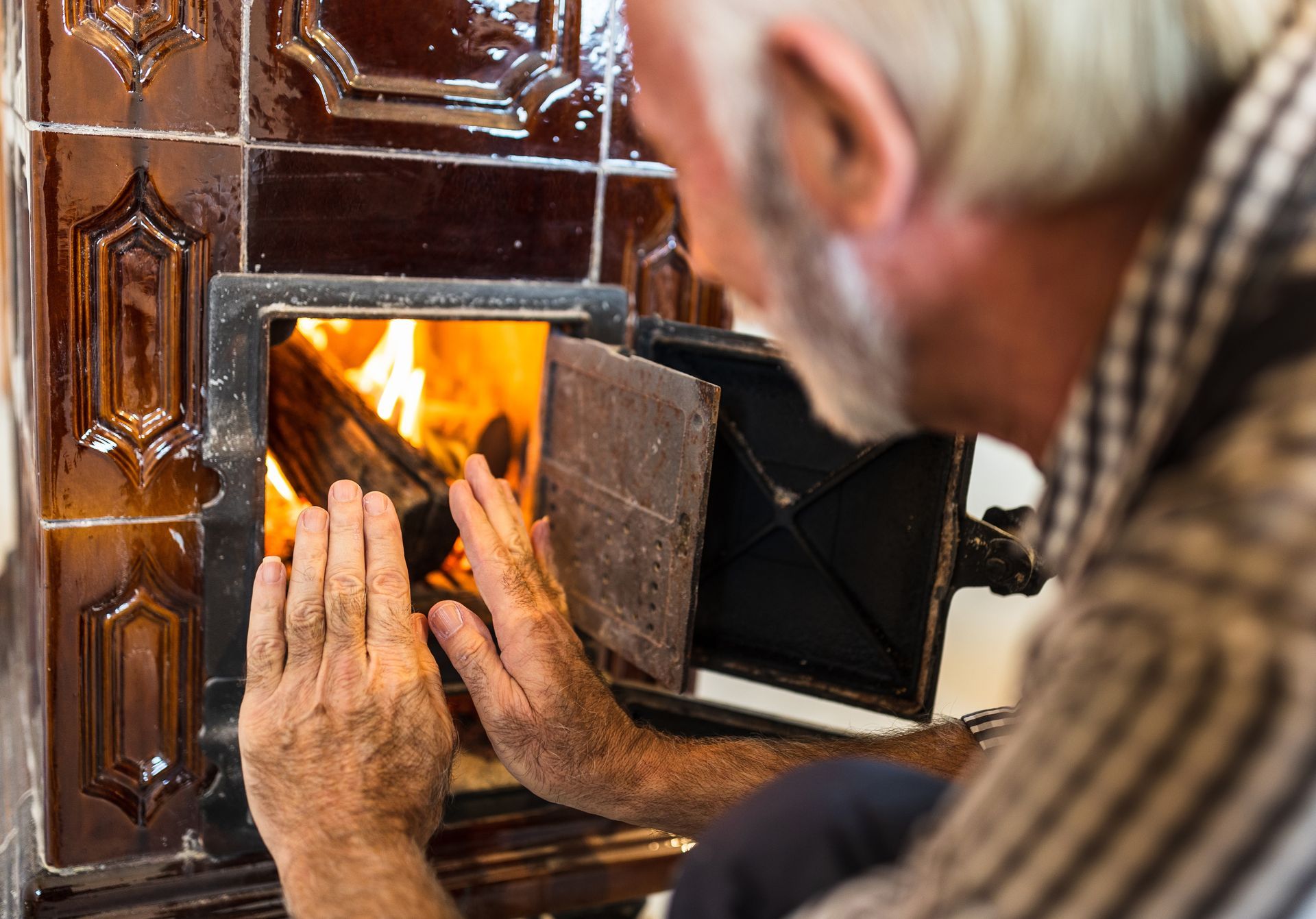 Man with gray beard tending a fire in a tiled stove, reaching out to feel the heat.