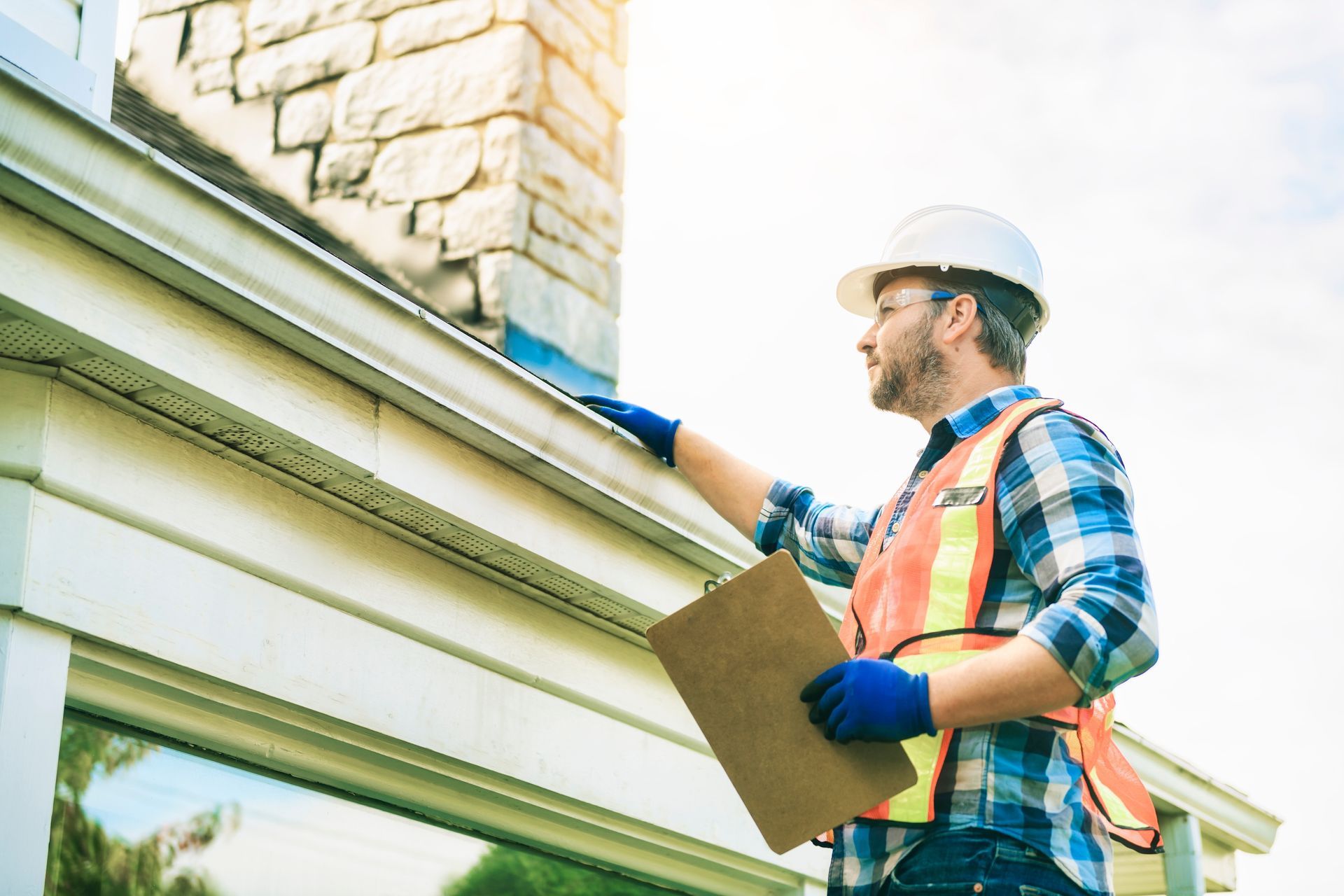 A man in a hard hat inspects a house gutter, holding a clipboard, near a stone chimney.