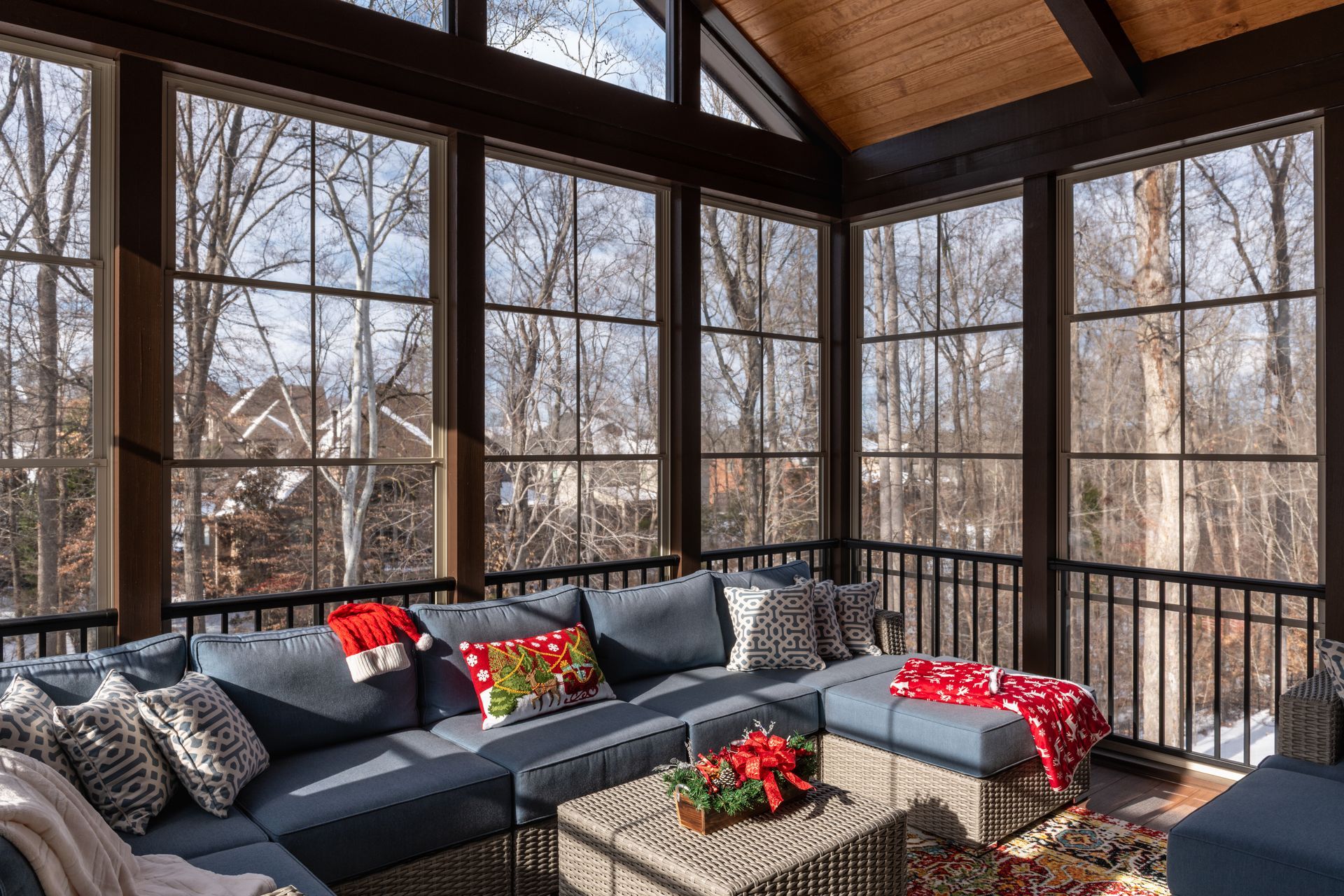 Cozy sunroom with large windows, blue sectional sofa, and holiday decorations. Winter trees visible outside.