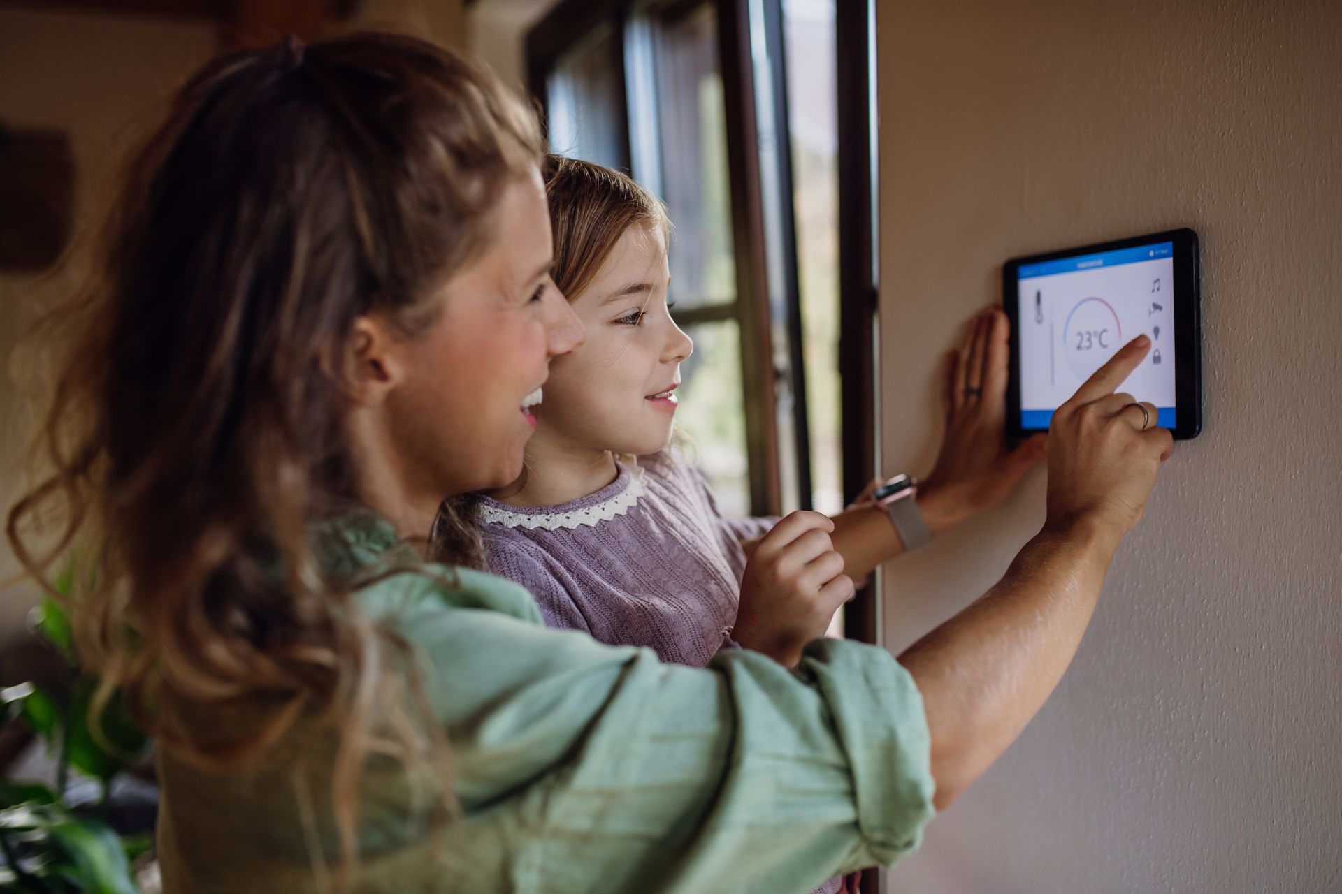 Woman and child use a touchscreen smart home control panel on a wall. The woman points, smiling.