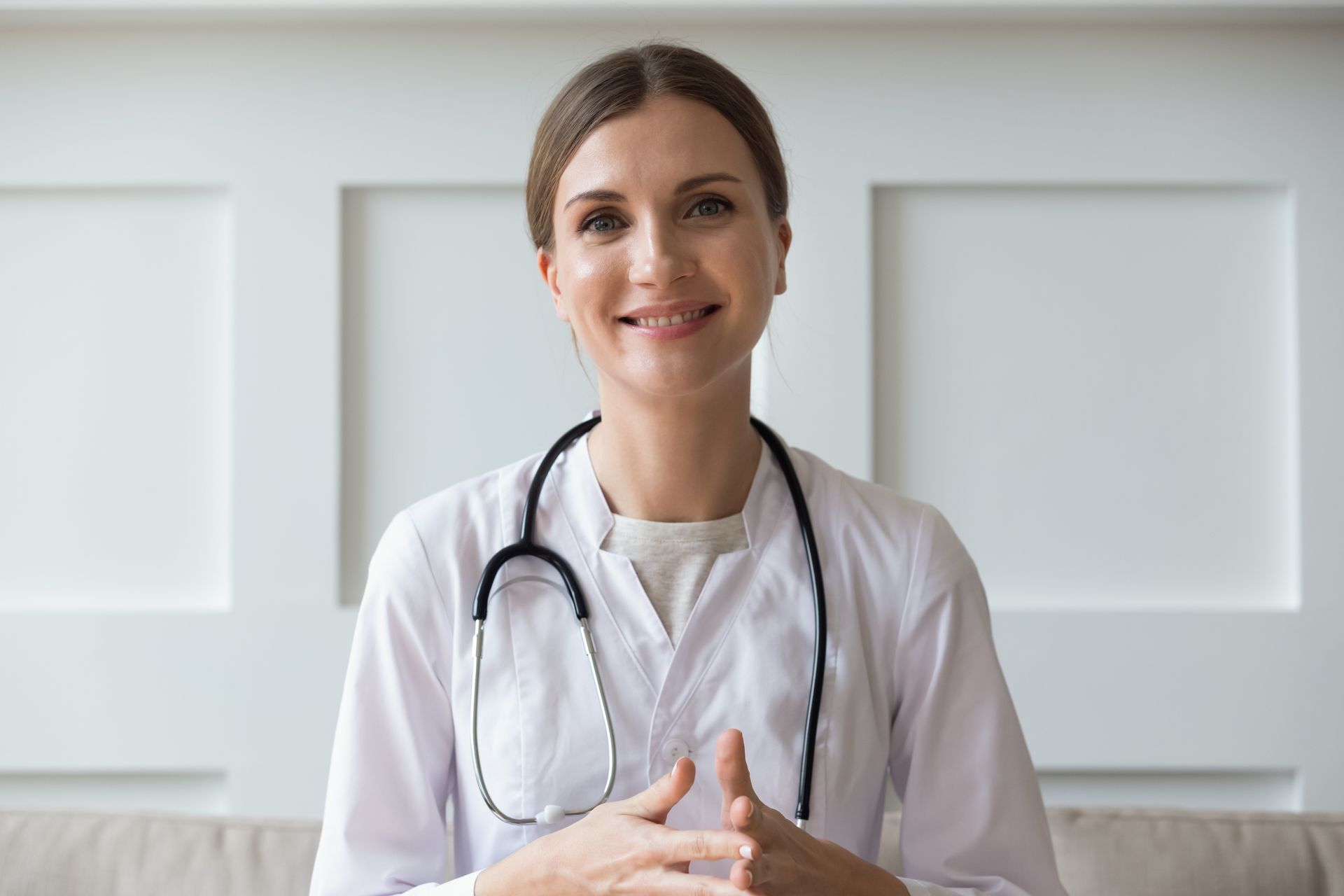 A female doctor with a stethoscope around her neck is smiling while sitting on a couch.