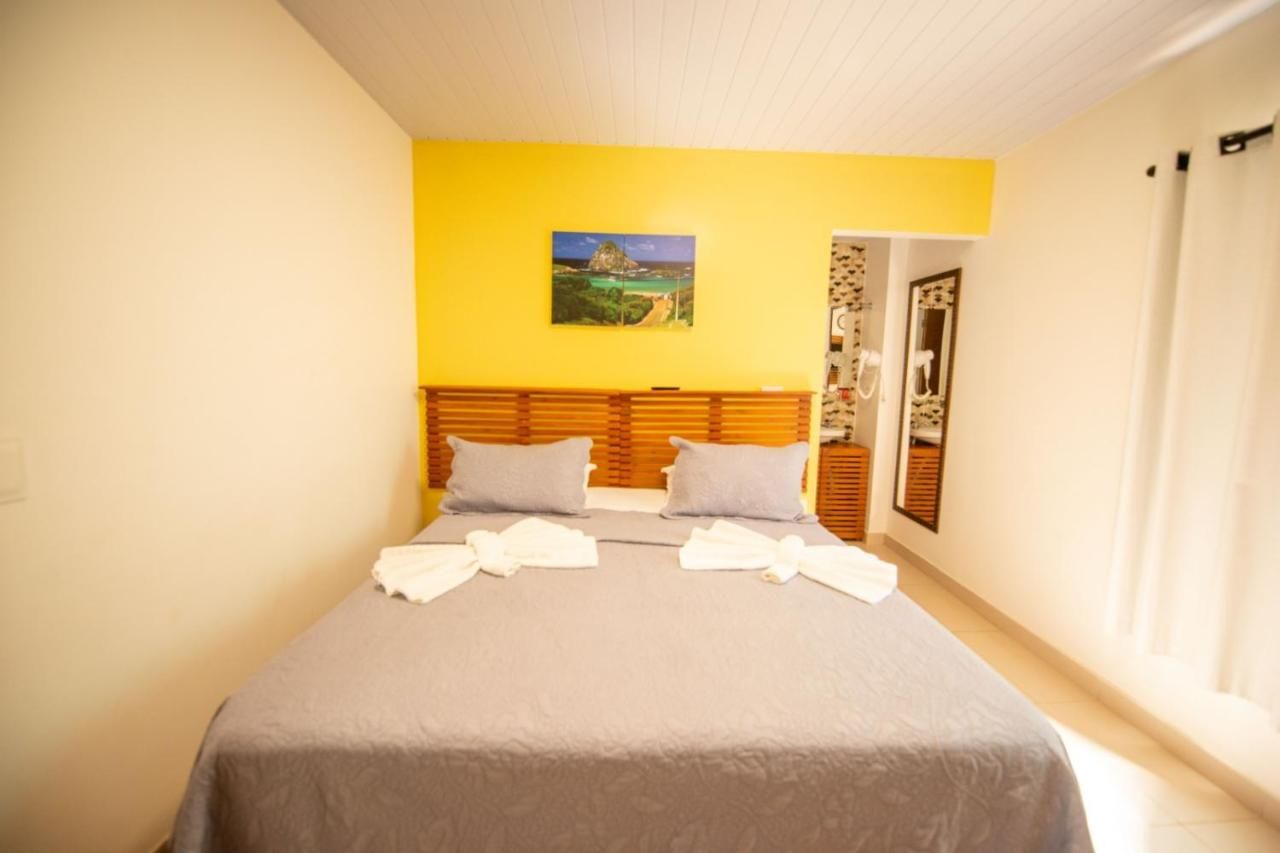 Quarto com cama cinza, parede com detalhes em amarelo, obras de arte e uma porta que dá acesso a outro ambiente com espelho.