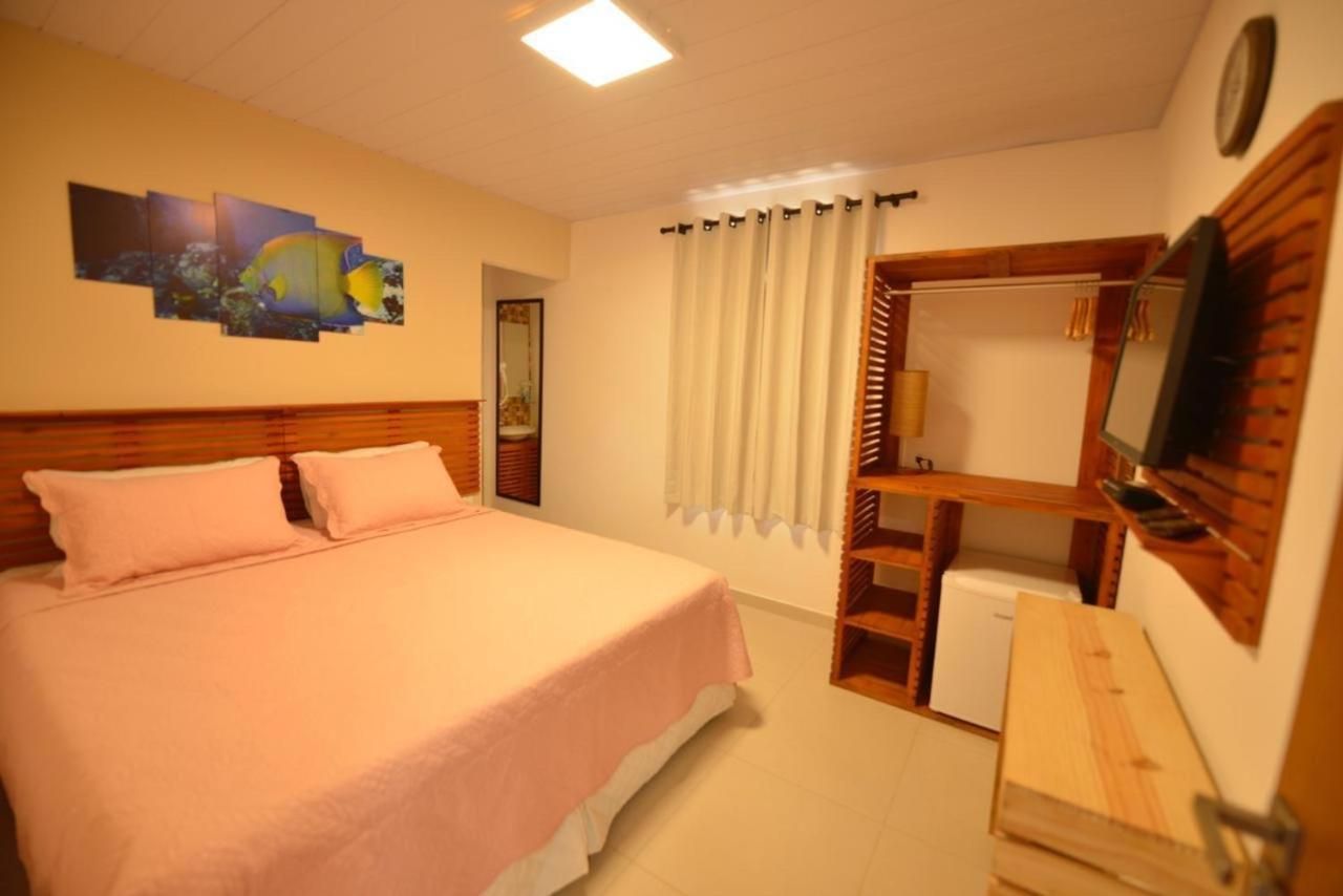 Quarto com cama, armário, TV e frigobar; roupa de cama salmão; detalhes em madeira.