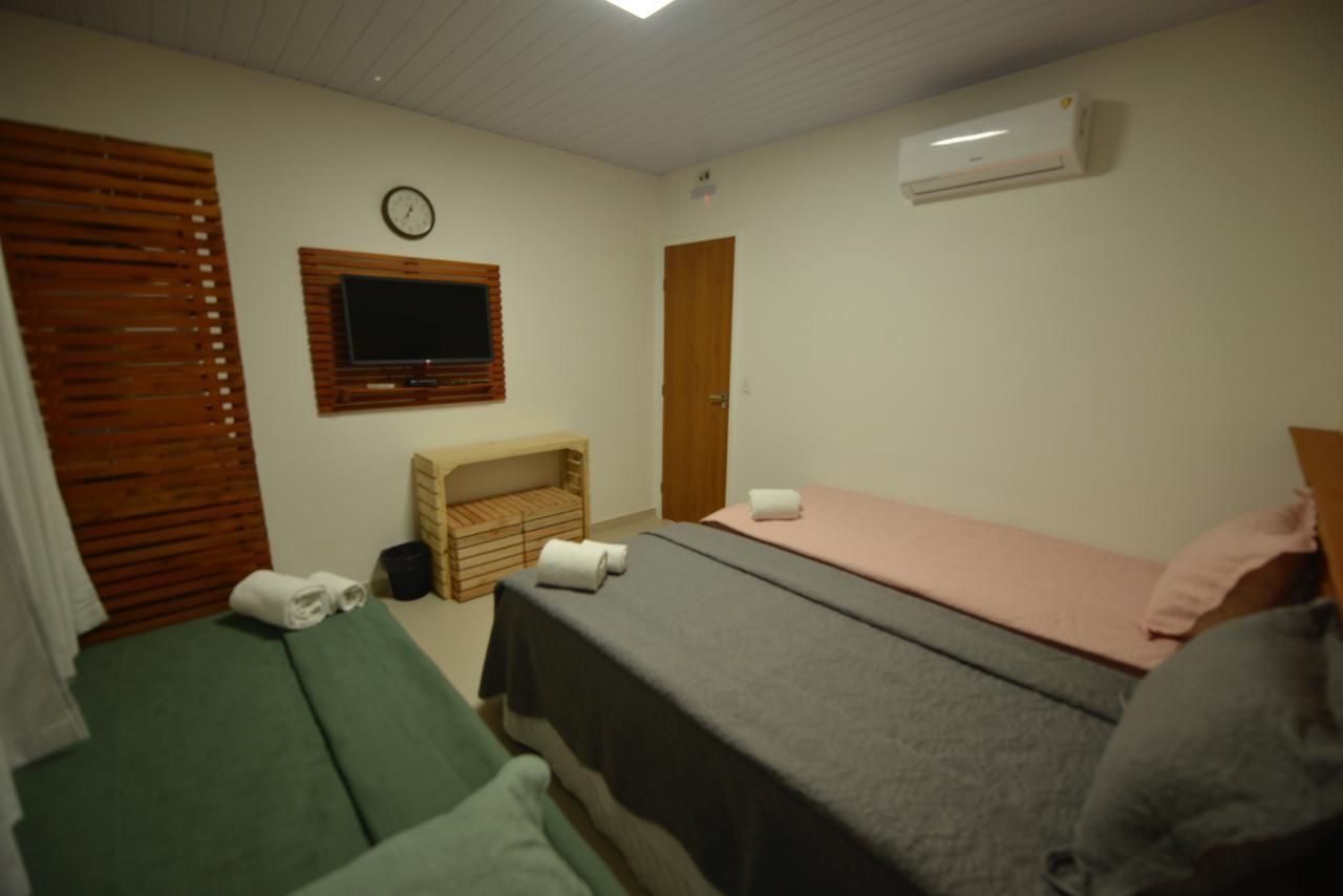 Quarto com duas camas, TV e ar condicionado. Paredes brancas, detalhes em madeira e toalhas nas camas.