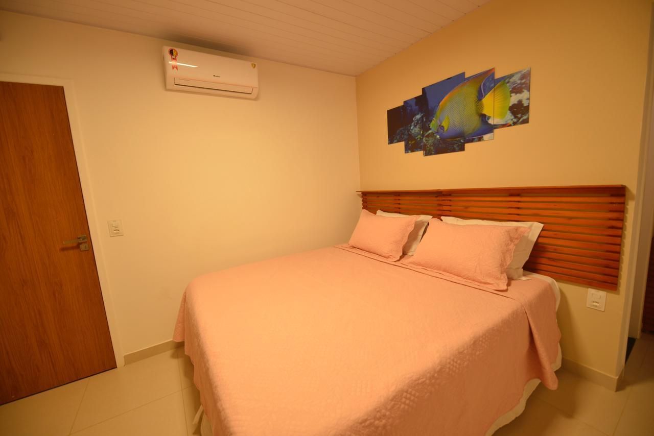 Quarto com cama, roupa de cama rosa, cabeceira de madeira, obras de arte, ar condicionado e porta fechada.