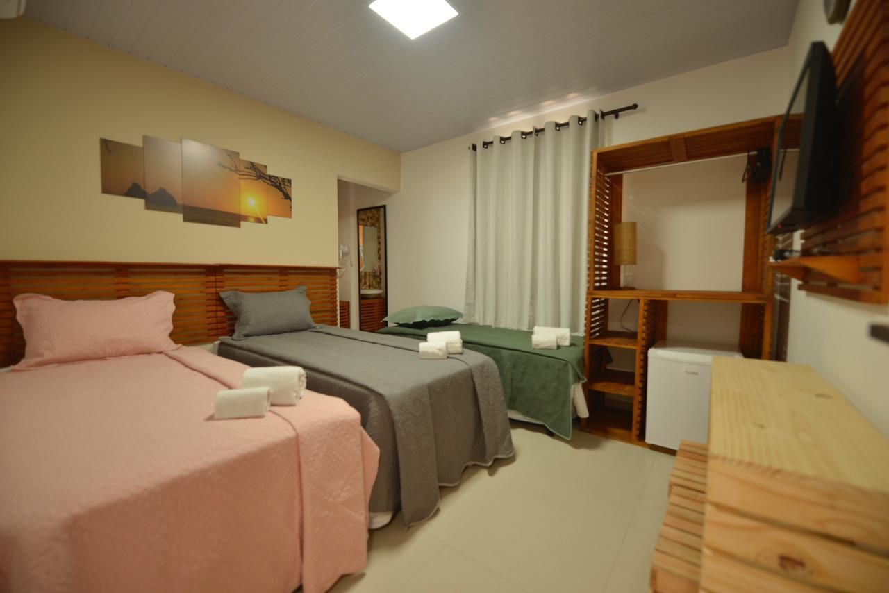 Quarto de hotel com três camas, roupa de cama em tons pastel, cabeceiras de madeira, frigobar e uma obra de arte com tema de pôr do sol.