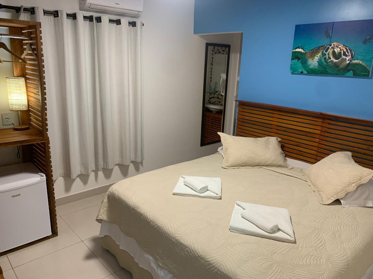 Quarto com cama, parede azul com desenho de tartaruga, cortinas e frigobar.