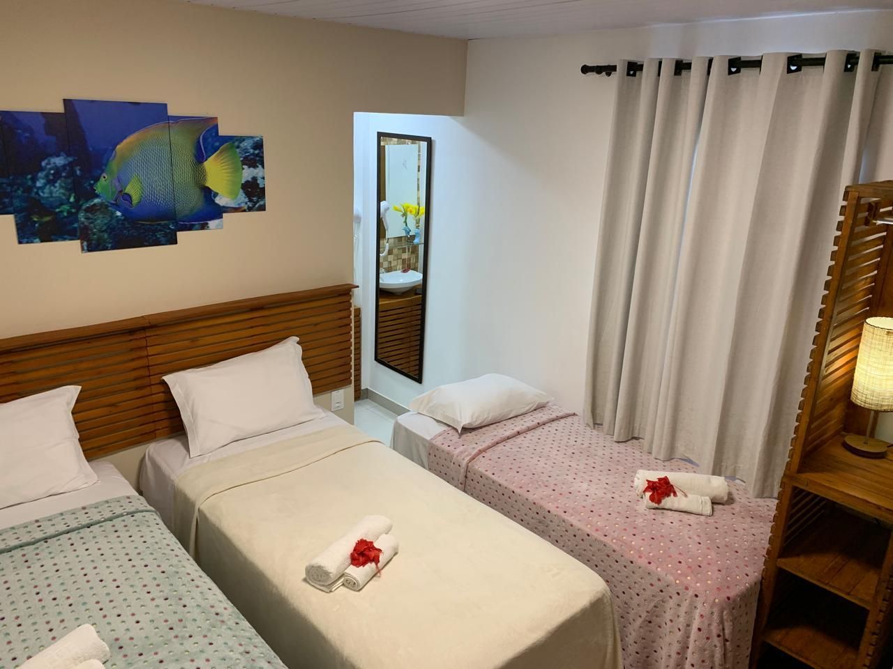 Quarto de hotel com três camas, decoração com tema marítimo, espelho e toalhas brancas. Colchas bege, brancas e rosa.