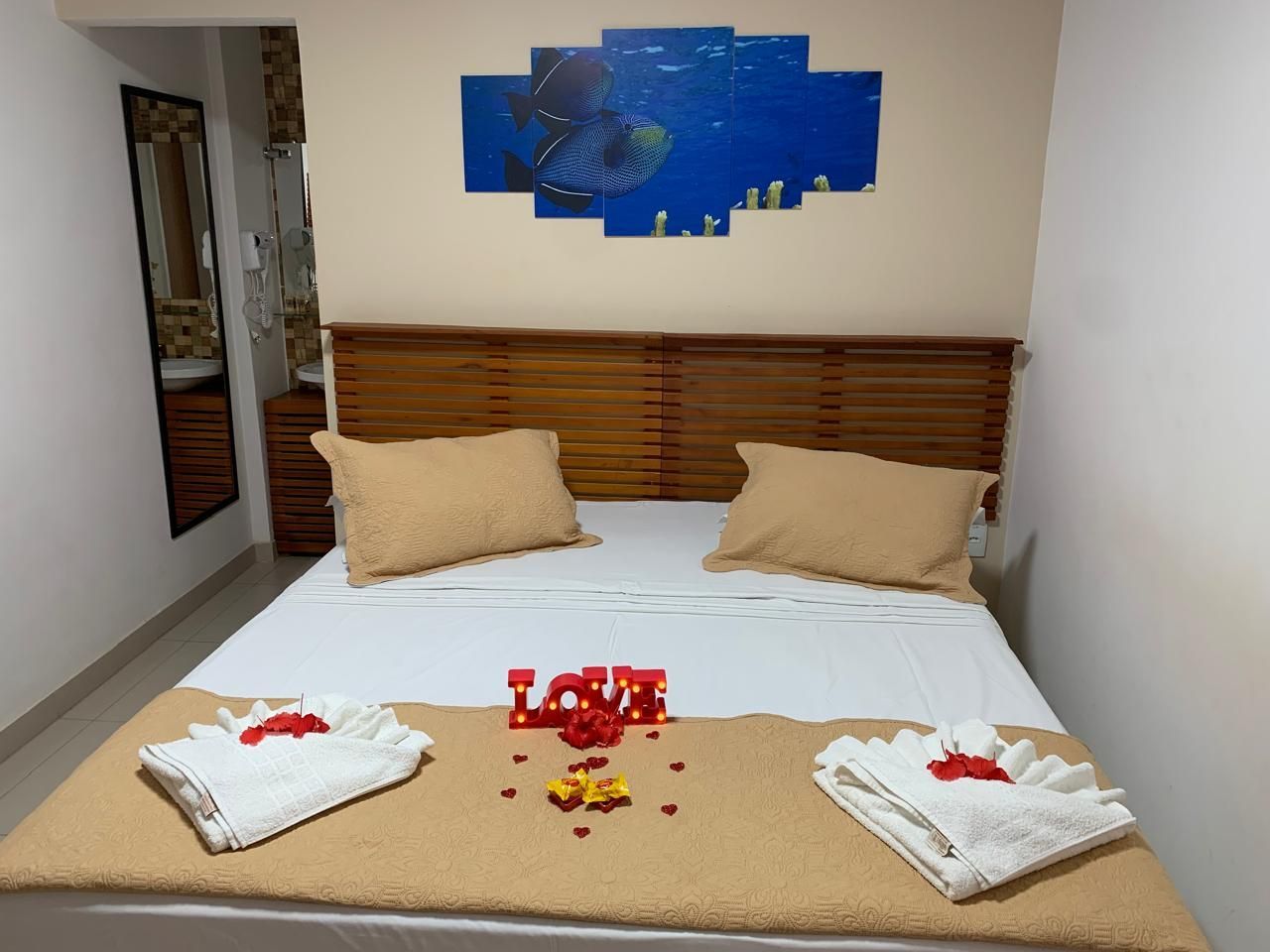 Quarto com cama arrumada, cabeceira, quadros na parede, toalhas e decoração.