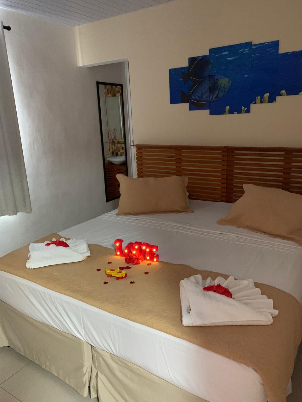 Quarto com cama arrumada, cobertor e travesseiros bege, toalhas dobradas e luzes decorativas vermelhas.