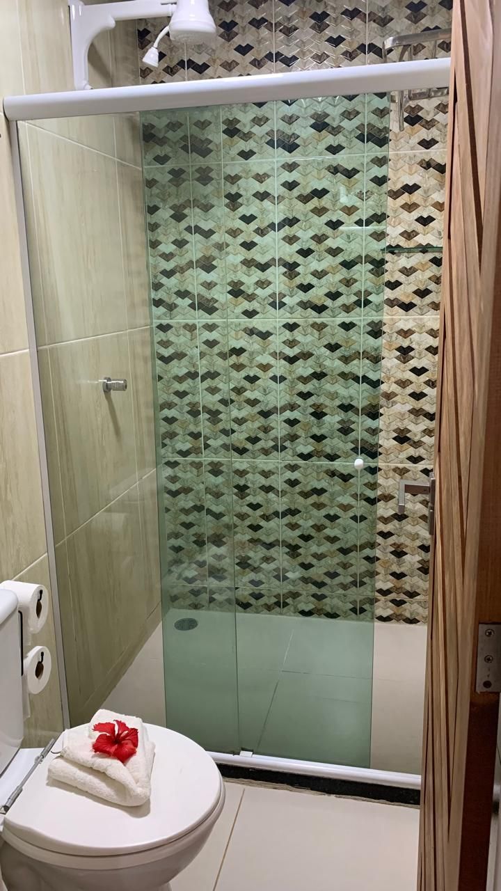 Banheiro com box de vidro, parede com azulejos estampados, vaso sanitário com toalhas dobradas e flor vermelha.