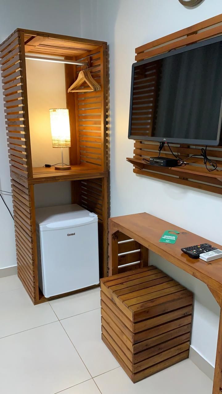 Armário e escrivaninha de madeira com frigobar, luminária, TV e banqueta em um quarto de hotel.