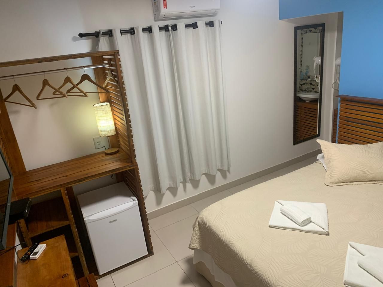 Um quarto de hotel com cama, guarda-roupa, frigobar e janela com cortinas.