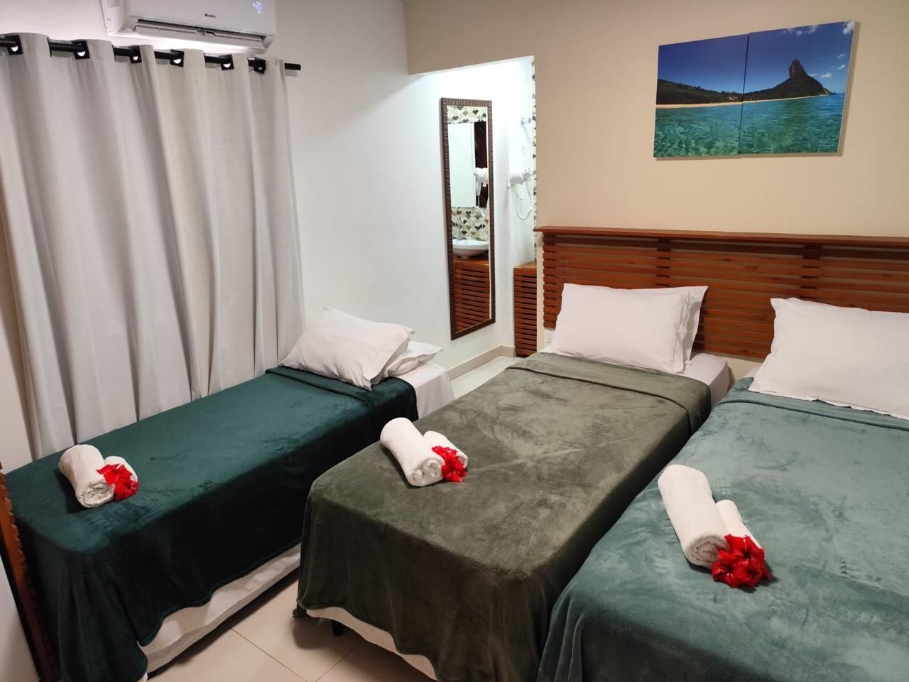 Três camas em um quarto de hotel com cobertores e toalhas verdes e cinzas, e uma pintura de paisagem.