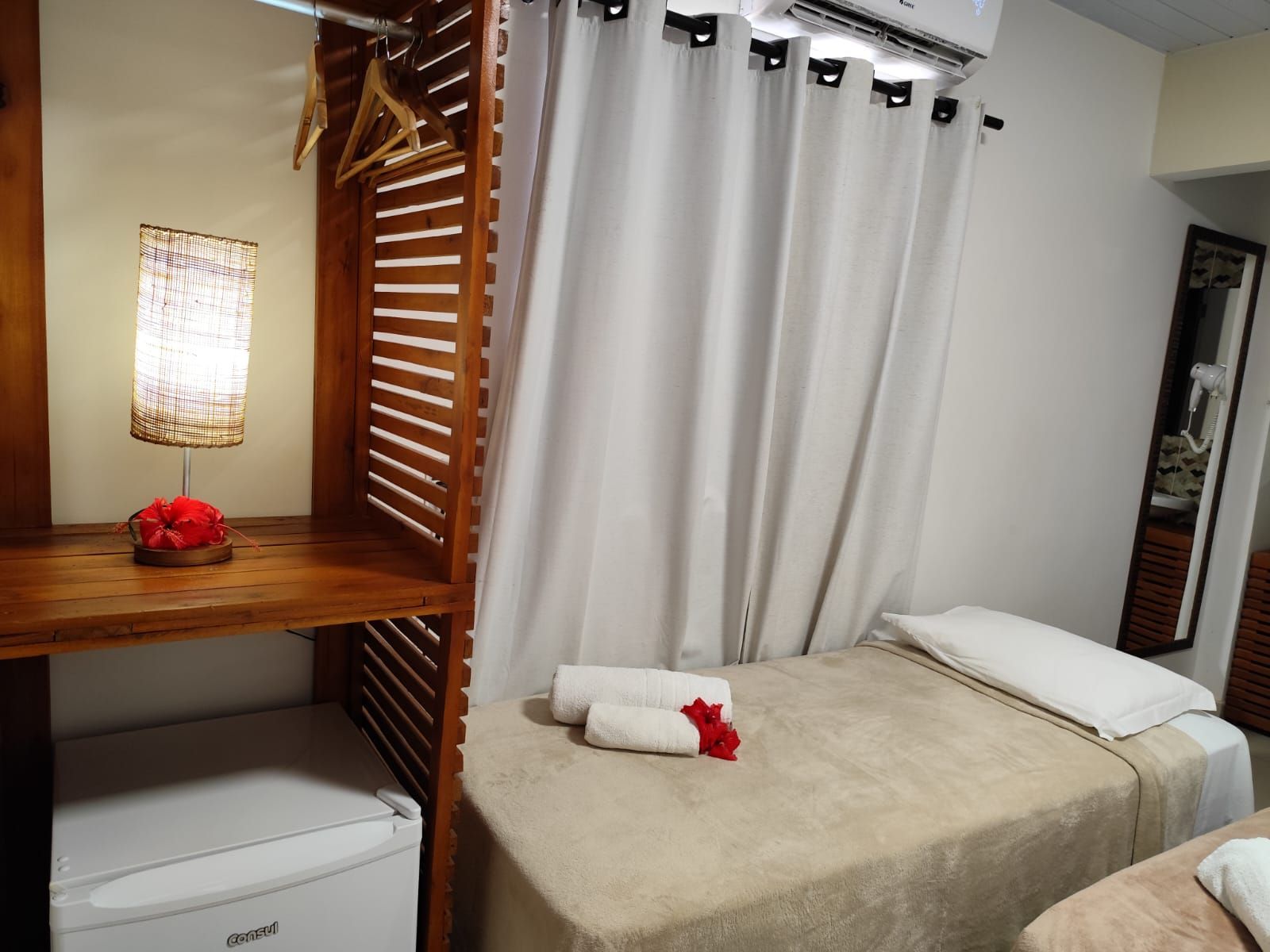 Quarto com duas camas, cortinas brancas, guarda-roupa de madeira, frigobar e uma flor vermelha decorativa.