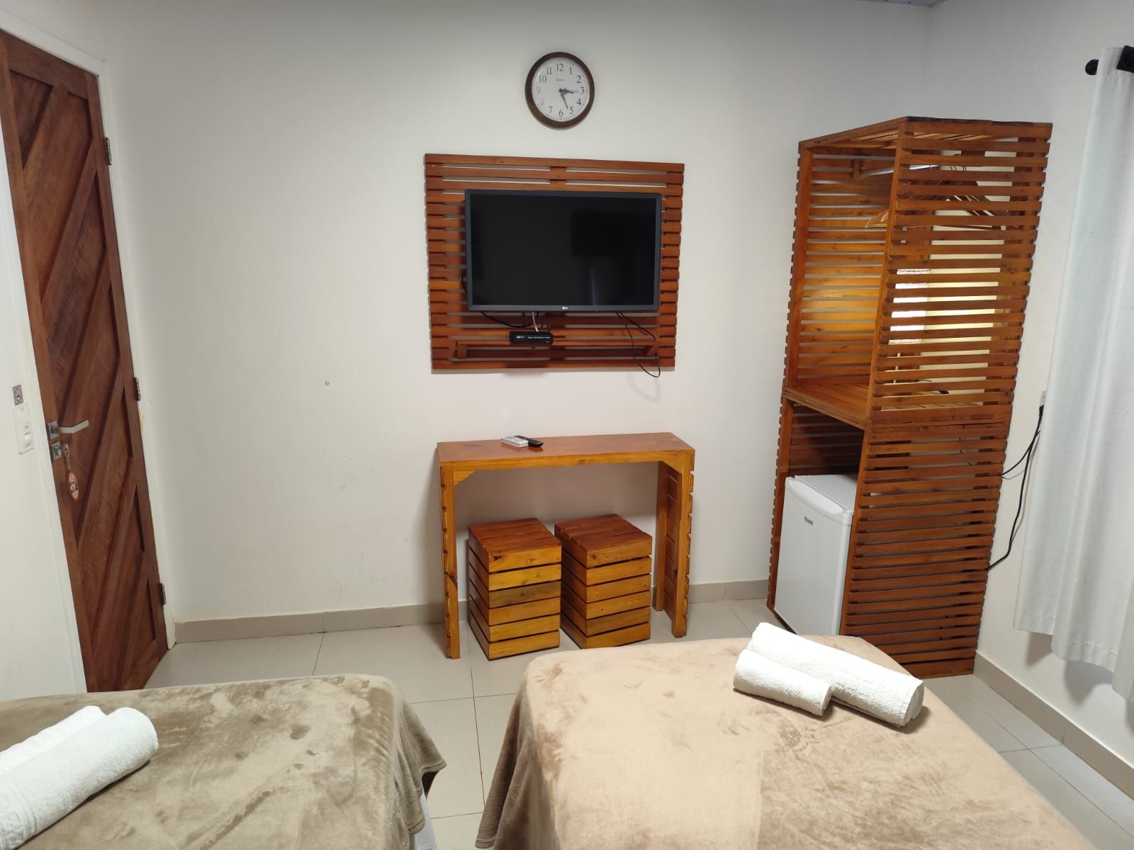 Quarto com duas camas, TV, móveis de madeira, relógio e frigobar. Paredes brancas e piso claro.