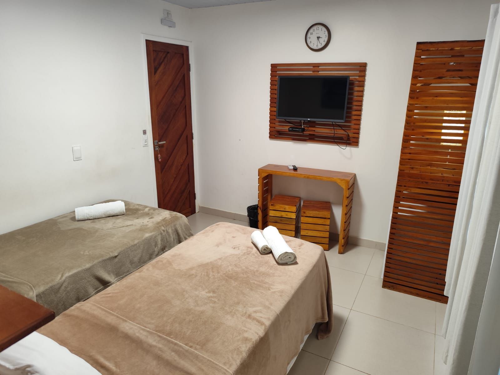 Quarto com duas camas de solteiro, TV, detalhes em madeira e relógio.